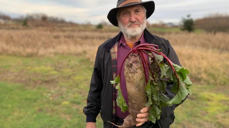 an older man holding a large root in his right hand and wearing a hat on top of it