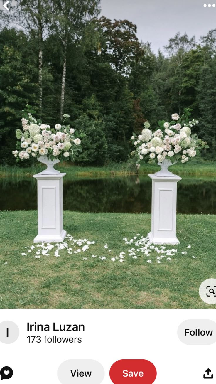 two white vases with flowers on them sitting in the grass near some water and trees