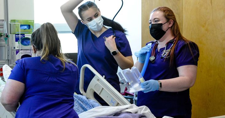 two women in scrubs and face masks standing around a hospital bed