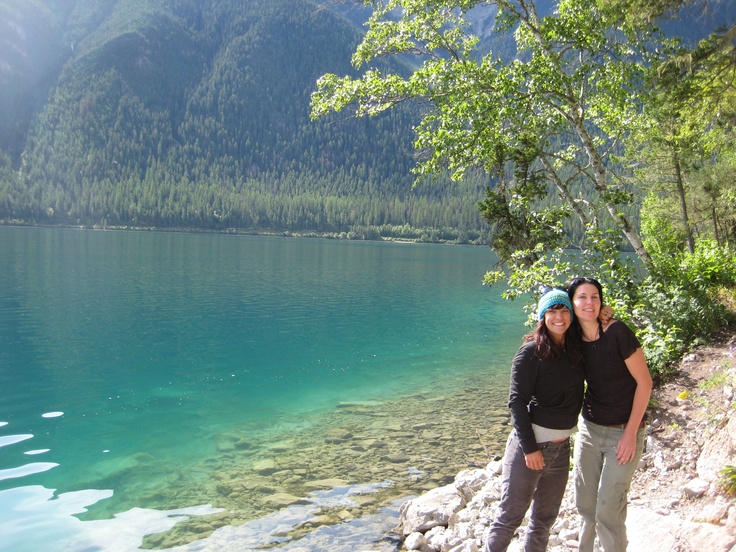 two women standing next to each other on the shore of a lake with mountains in the background