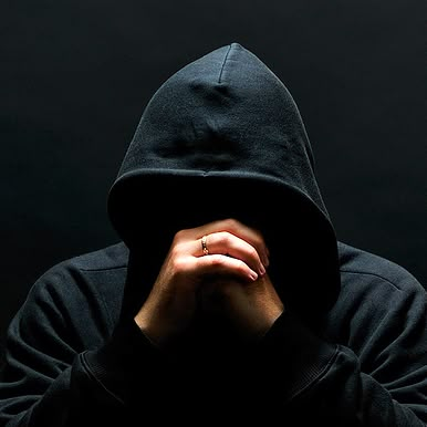 a man in a black hoodie is covering his face with his hands while standing against a dark background