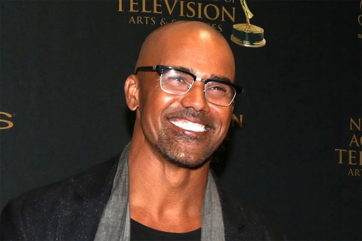 a bald man with glasses and a scarf around his neck smiles at the camera while standing in front of a black backdrop