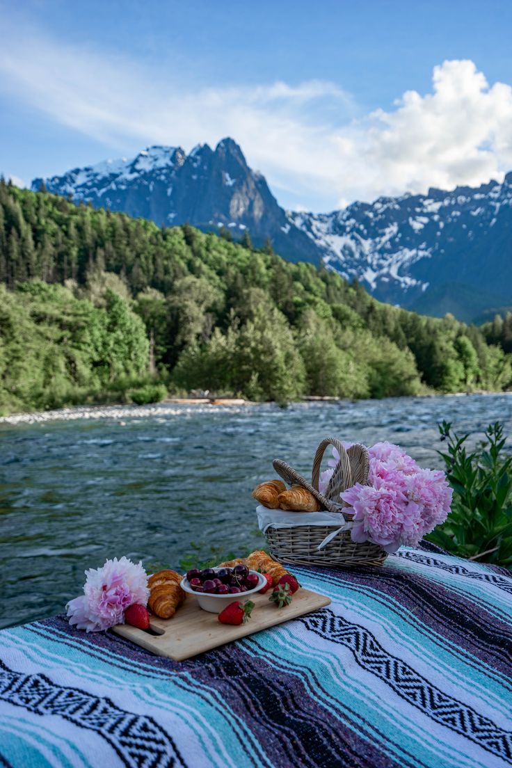 food and flowers on a picnic table overlooking a river with mountains in the backgroud