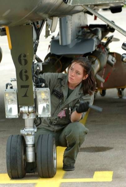 a woman in uniform sitting on the ground next to an air force jet engine and looking at the camera