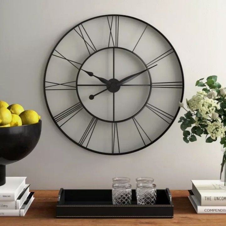 a large clock on the wall above a bowl of lemons and some books next to a vase with flowers