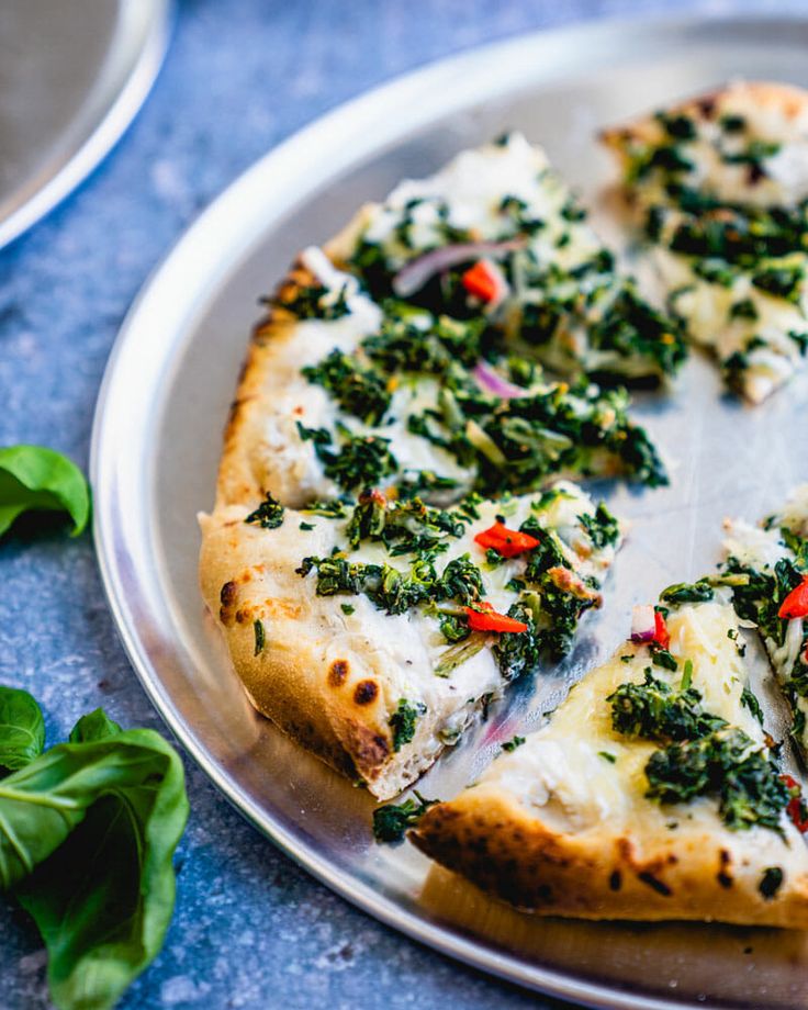 a pizza with spinach and cheese is on a silver plate next to some basil leaves