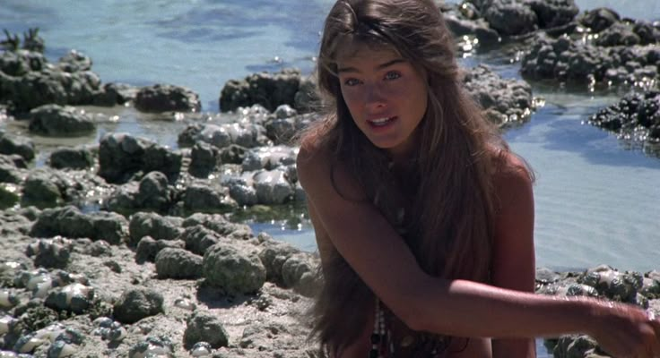 a young woman sitting on the beach next to some rocks and seaweed in her hair