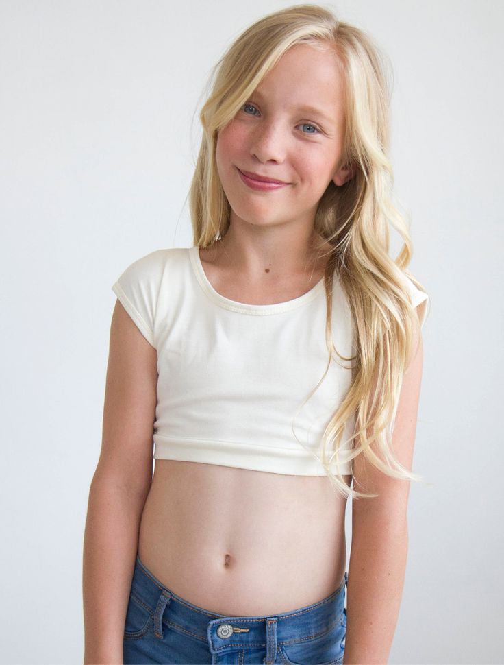 a young blonde girl with no shirt on posing for the camera, wearing blue jeans and a white crop top