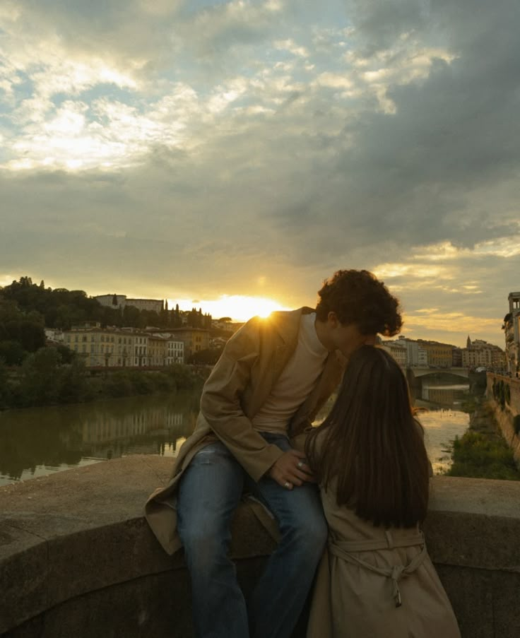 a man and woman are kissing on a bridge over the water at sunset with buildings in the background