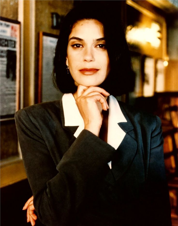 a woman wearing a suit and tie standing in front of a bar with her hand on her chin