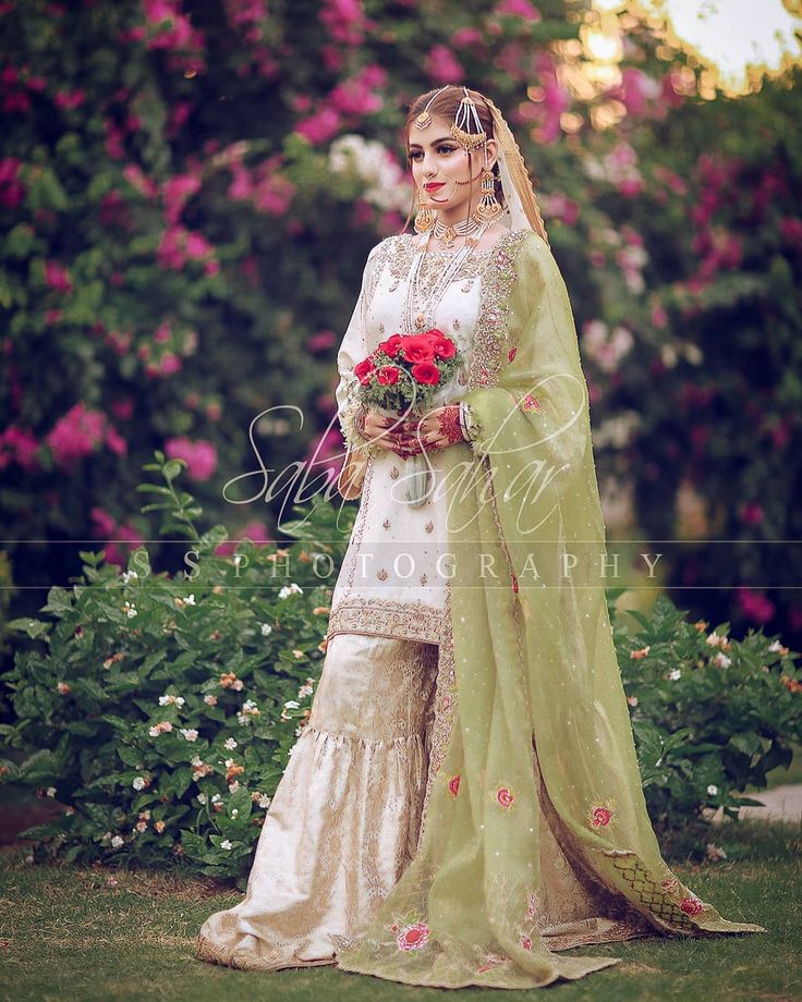 a woman in a white and green wedding outfit holding a bouquet of flowers, posing for the camera