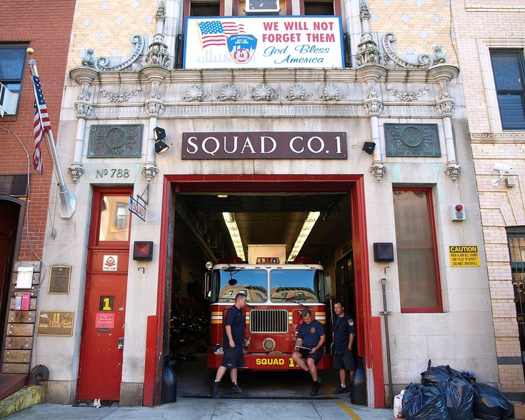 two men standing in front of a fire station with a truck parked outside the door