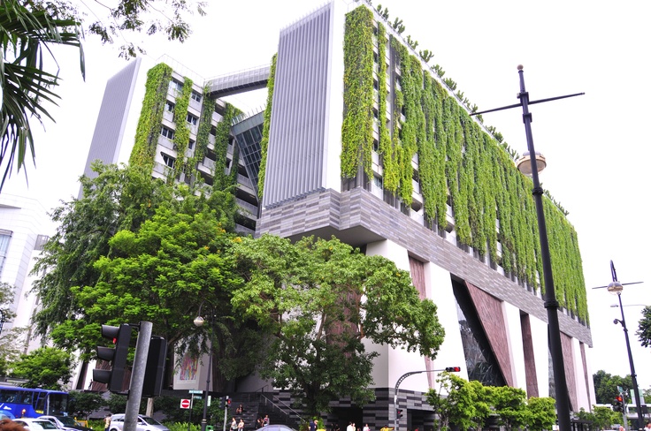 the building is covered in green plants and has people walking on the sidewalk near it