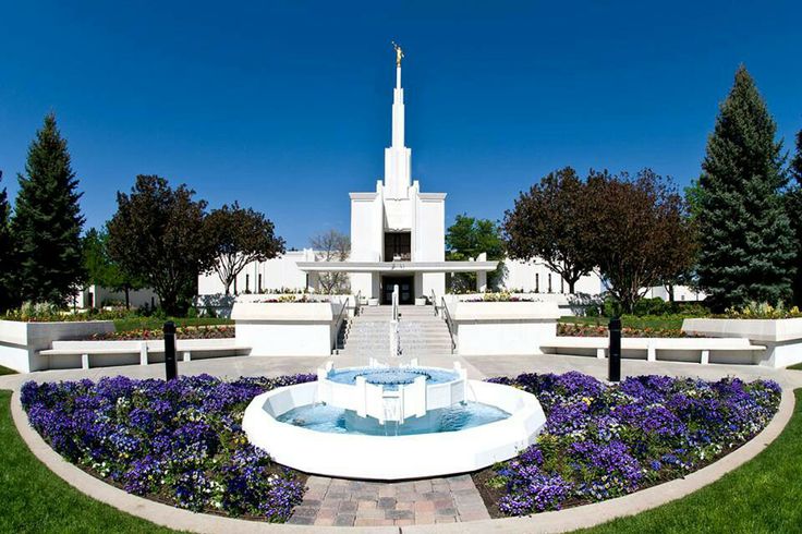 a fountain in front of a church with purple flowers on the ground and trees around it