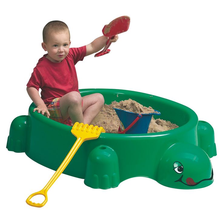 a little boy sitting in a green sand pit with a shovel and toy turtle on it