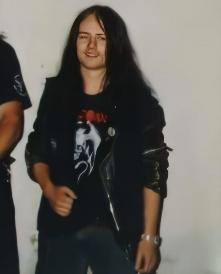 two men standing next to each other in front of a white wall and one is wearing a leather jacket