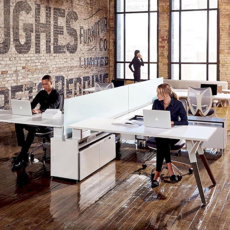 two people sitting at desks in an office with large windows and brick wall behind them