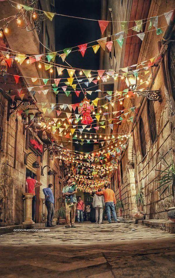 several people are walking down an alley way with colorful lights strung over the walkways