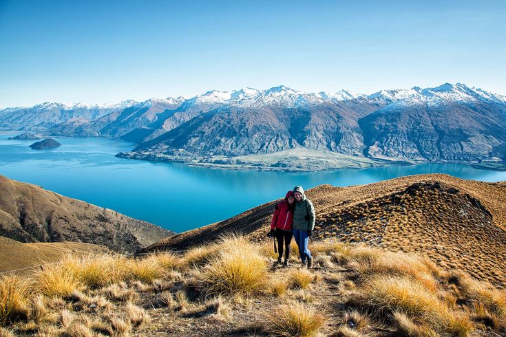 two people standing on the side of a mountain looking out over a lake and mountains