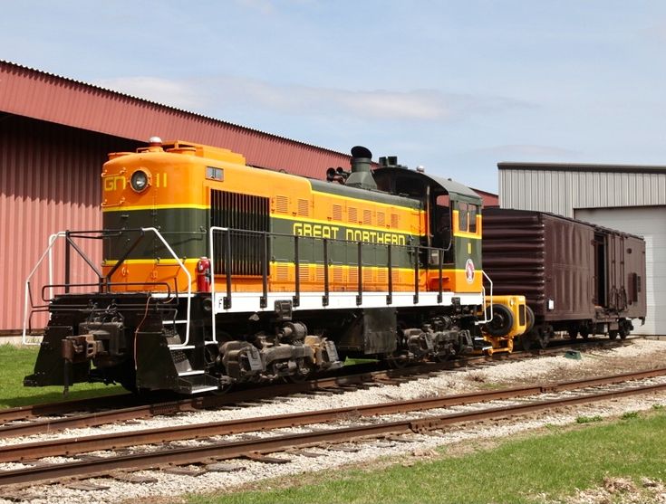 an orange and green train is on the tracks next to a red building with two doors