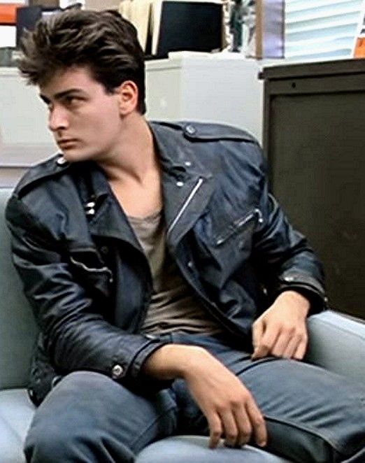 a young man sitting on top of a blue couch in an office setting, wearing a black leather jacket and jeans