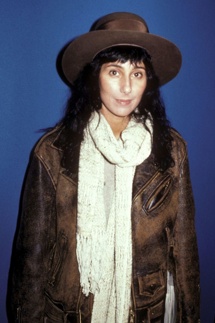 a woman wearing a hat and scarf standing in front of a blue wall with her hands on her hips