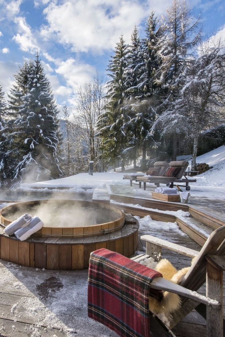 an outdoor hot tub surrounded by snow covered trees and benches with blankets on the ground