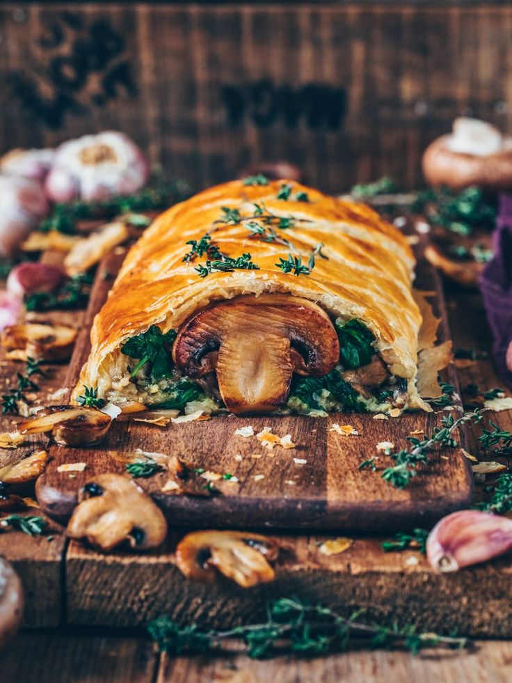 a wooden cutting board topped with a pastry covered in mushrooms and herbs next to other food items