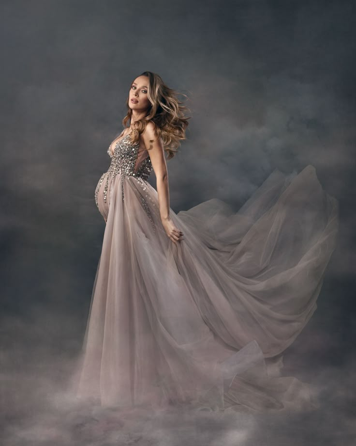 a woman in a long gray dress standing on a cloud covered ground with her hair blowing in the wind