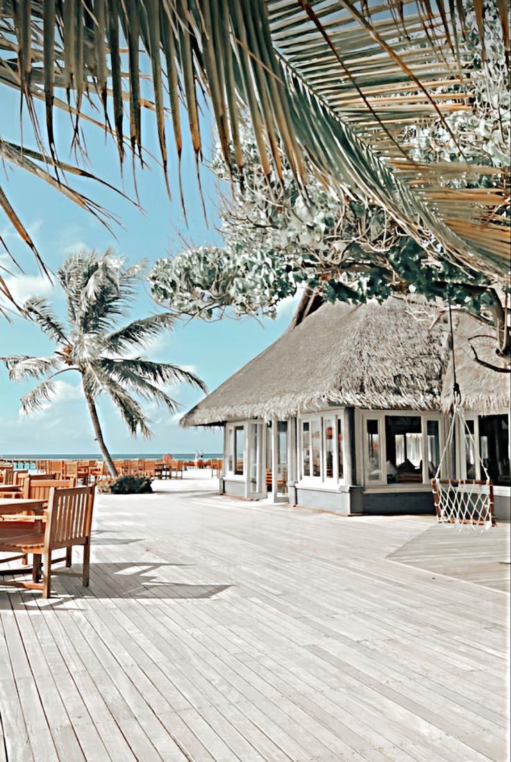 a wooden deck with tables and chairs under a thatched roof next to the ocean