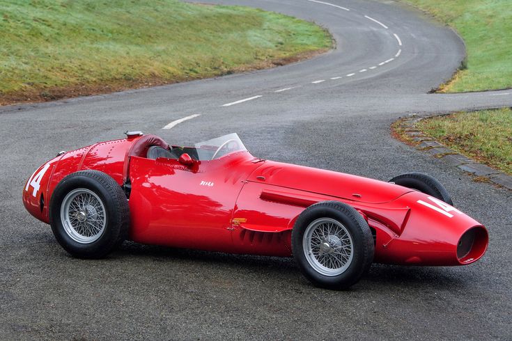 an old red race car driving down a winding road with grass and trees in the background