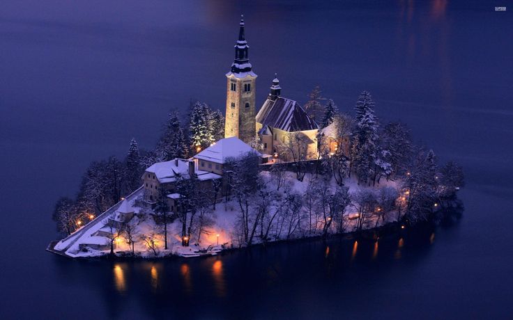 an island with a church lit up in the snow at night, surrounded by trees and water