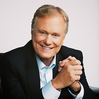 a smiling man in a black suit and blue shirt sitting on a chair with his hands folded