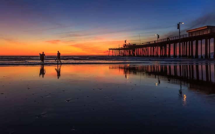 two people are standing on the beach at sunset with a pier in the back ground