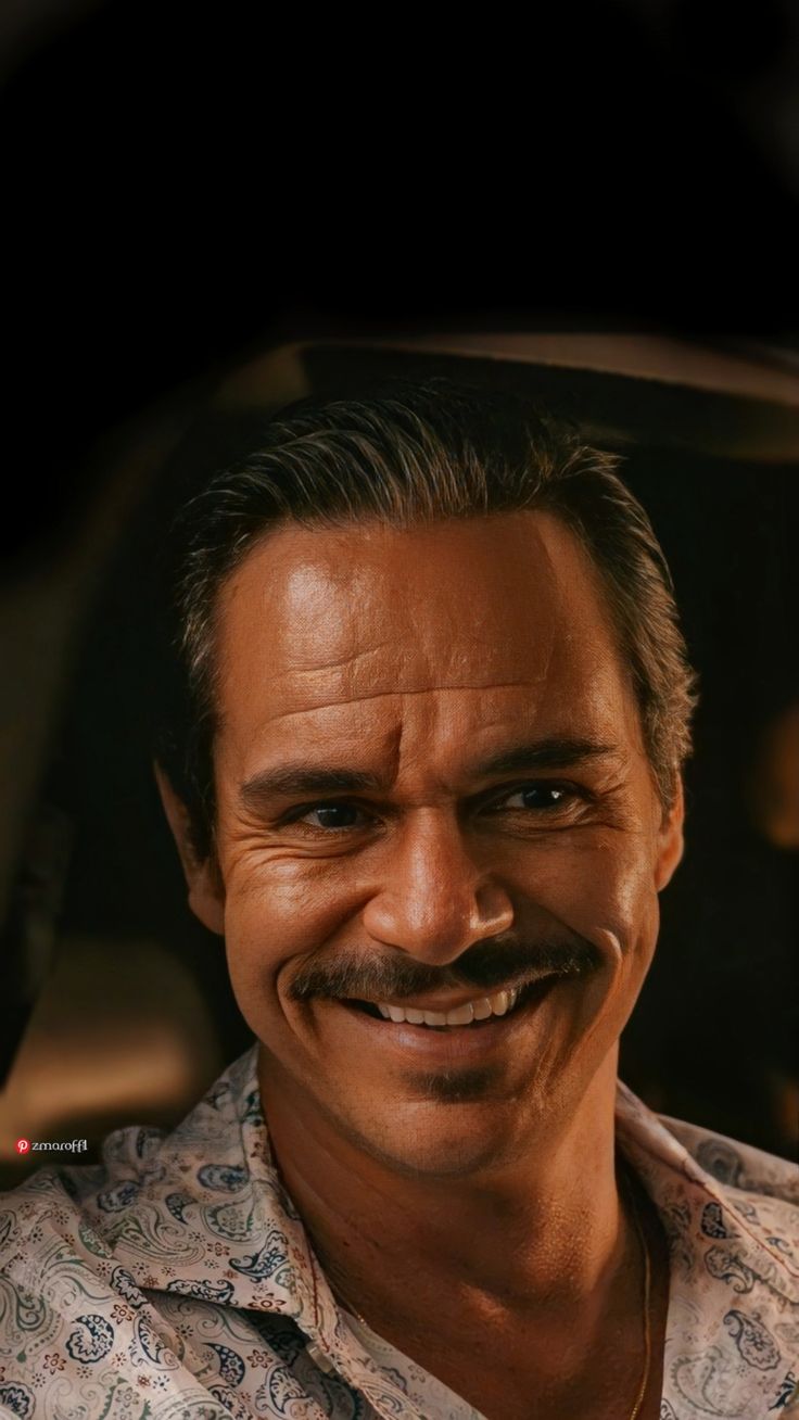 a man with a moustache smiling at the camera while sitting in a car