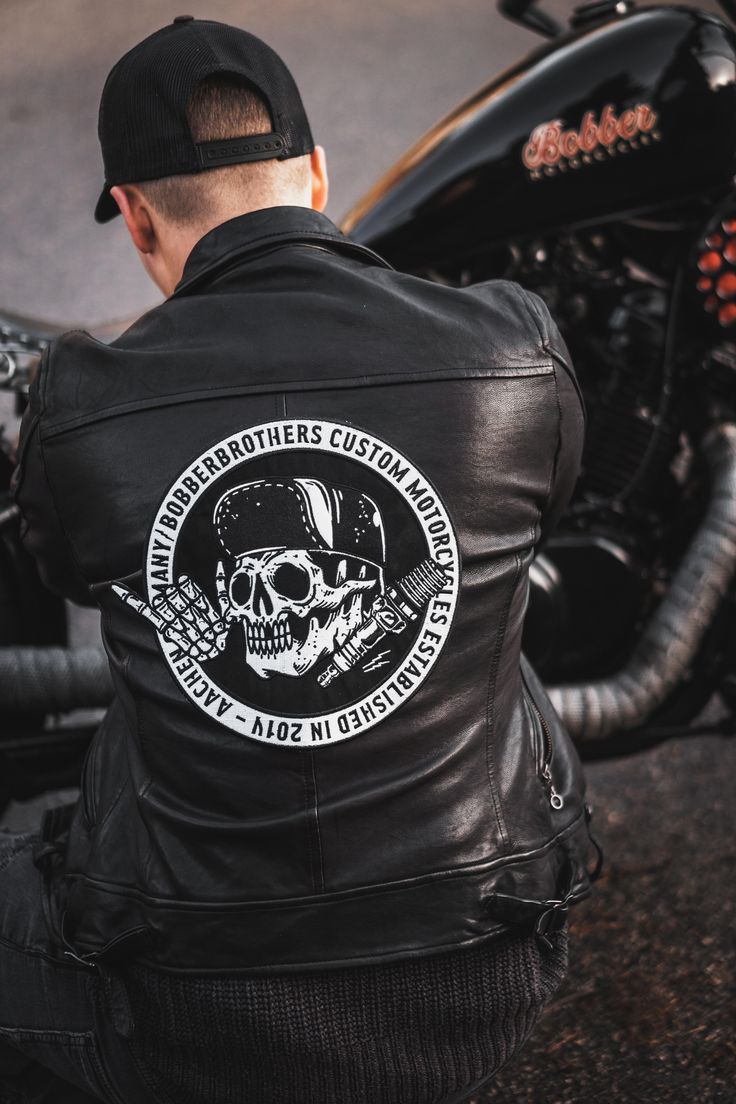 a man wearing a black leather jacket with a skull on it sitting next to a motorcycle