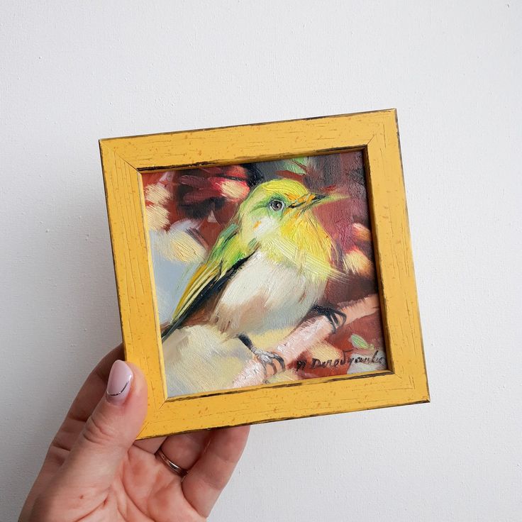 a painting of a bird in a yellow frame on a white wall with a hand holding it