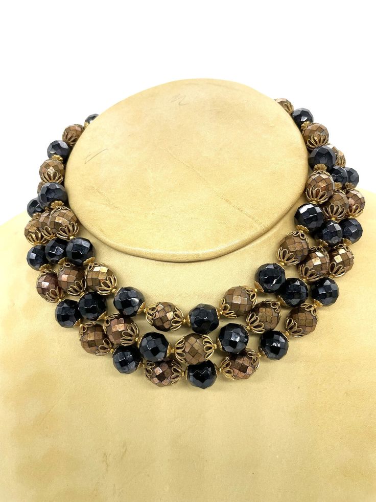 Embrace the vintage charm with this sophisticated triple-strand glass bead choker. Each strand, measuring 12 1/2 to 13 1/2 inches in length, is a testament to classic allure, featuring 11 millimeters faceted black beads and 10mm bronze glass beads. The bronze beads are accentuated with ornate gold-colored filigree bead caps, adding a touch of intricate detail to the necklace. An added 2-inch extension chain ensures a comfortable and adjustable fit. This choker is a striking blend of retro elegan Bead Choker, Gold Filigree, Glass Gifts, Beaded Choker, Bead Caps, Black Beads, Glass Bead, Vintage Charms, The Vintage