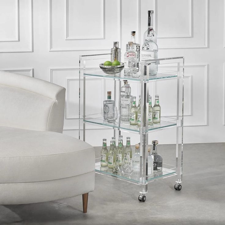 a clear bar cart with bottles and glasses on it next to a white chair in a room