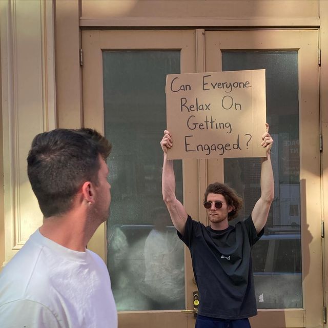 a man holding up a sign that says can everyone relax on getting engaged? with another man standing next to him