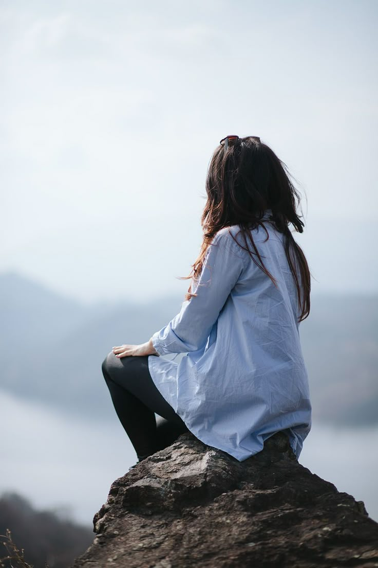 a woman sitting on top of a rock looking out over the water and mountains in the distance