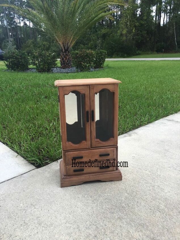 a wooden cabinet sitting on top of a sidewalk next to a lush green field and palm tree