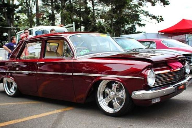 an old red car parked in a parking lot next to other antique cars and people