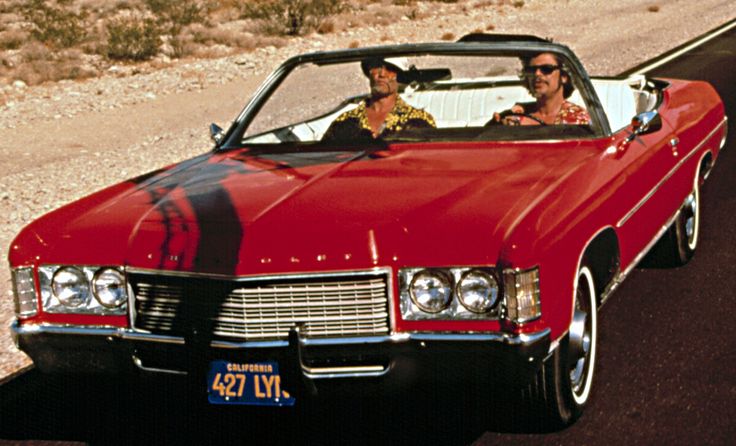 two people in a red convertible car driving down the road next to a desert landscape