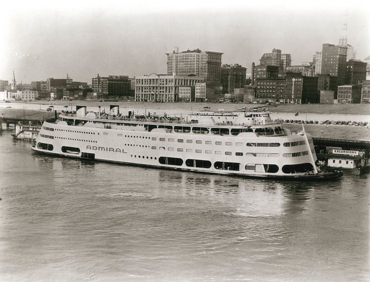 a large white boat floating on top of a river next to a city filled with tall buildings