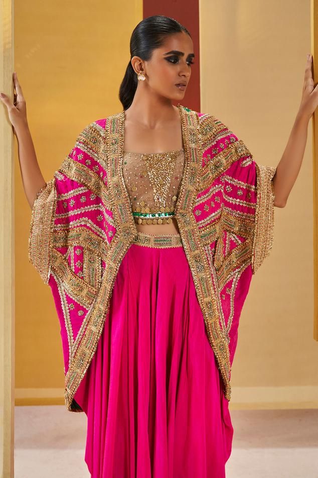 Fuchsia dupion silk cape with gota, fusion thread and zardosi embroidery. Comes with draped sharara pant and a net blouse.
Components: 3
Pattern: Embroidered
Type Of Work: Gota, Fusion thread, Zardosi
Neckline: Square Neck
Sleeve Type: Blouse : Sleeveless, Cape : Flared Sleeves
Fabric: Dupion silk, Blouse : Net
Color: Fuchsia
Other Details: 
Coin tassels
Note : Outfit worn by the model on the right is not for sale.
Occasion: Mehendi and Haldi - Aza Fashions Bollywood Style Choli With Cape Sleeves And Cutdana, Anarkali Choli With Cape Sleeves, Pink Bollywood Kaftan With Dupatta, Bollywood Style Pink Saree Kaftan, Bollywood Style Sharara With Cape Sleeves For Festivals, Designer Pink Kaftan With Traditional Drape, Traditional Sharara With Gota Work And Cape Sleeves, Traditional Sharara With Cape Sleeves For Festivals, Pink Anarkali Kaftan For Festive Occasions