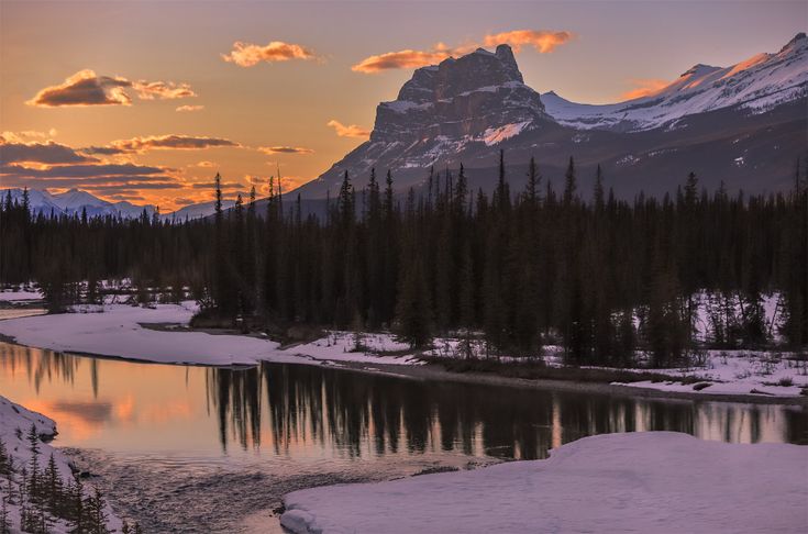 the sun is setting over a mountain range and a river in the foreground with snow on the ground