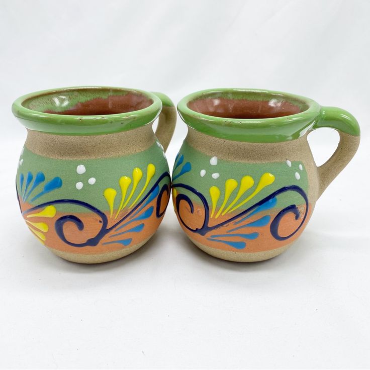 two ceramic mugs sitting side by side on a white surface with designs painted on them