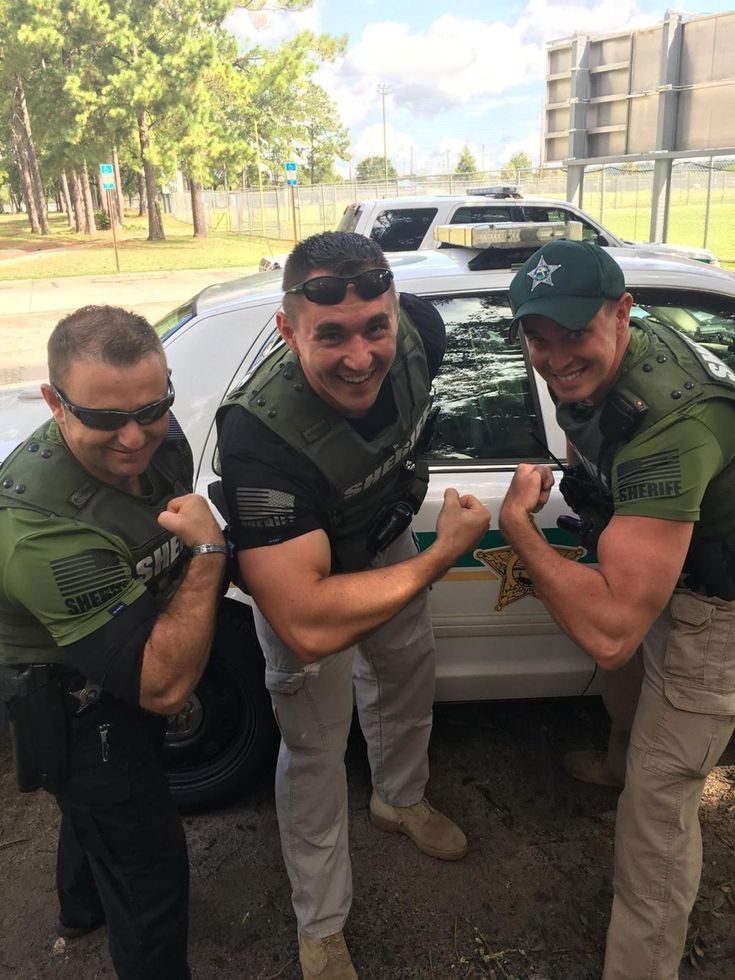 three men in police uniforms standing next to a white car with their arms wrapped around each other