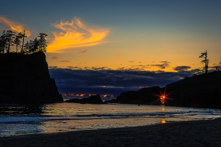 the sun is setting at the beach with some trees in the foreground and another rock outcropping on the shore